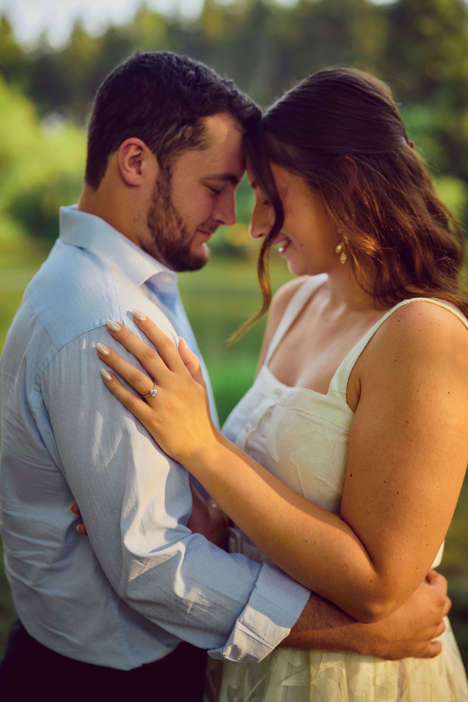 A romantic couple embraces outdoors in formal attire with a dreamy forest backdrop during golden hour.