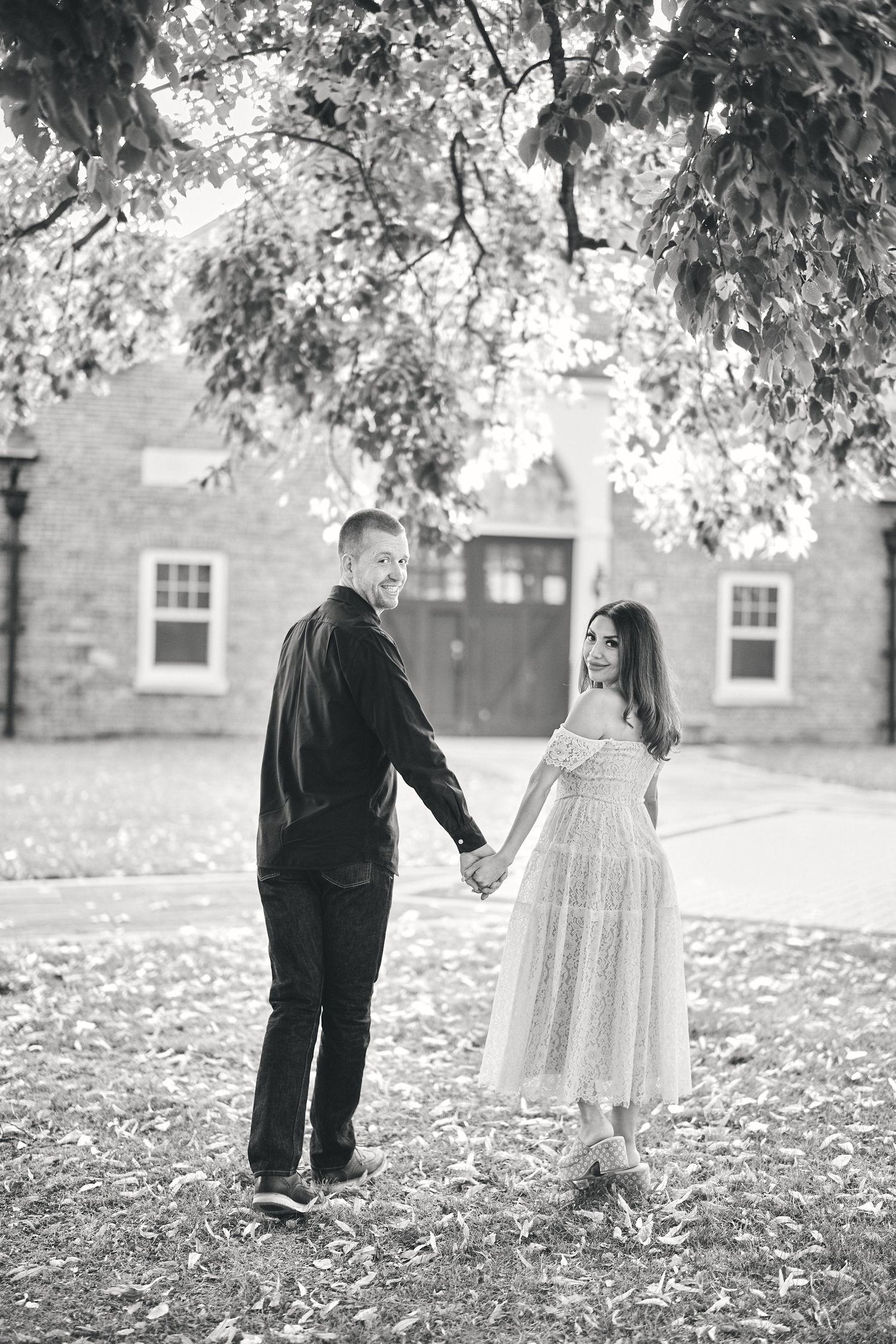 Black and white photo of couple holding hands in front of brick building with overhanging tree branches.