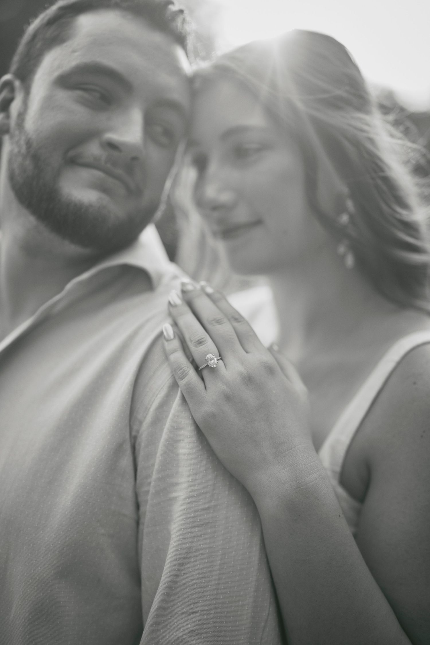 A black and white intimate engagement photo showing a couple embracing as sunlight illuminates a diamond ring.