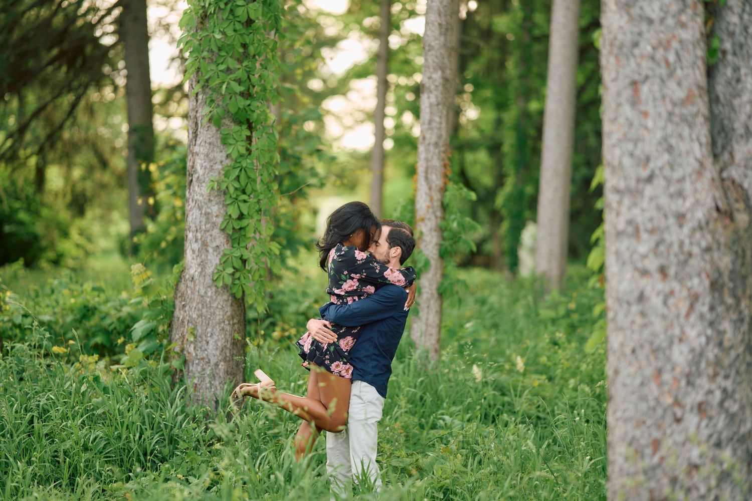 A loving embrace between two people standing in a serene forest setting surrounded by tall trees and lush greenery.