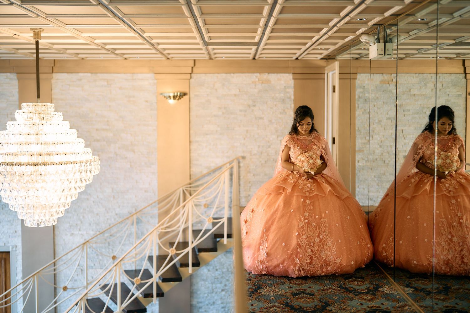 A person in a peach-colored ballgown stands near a crystal chandelier and mirrored wall in an elegant room.