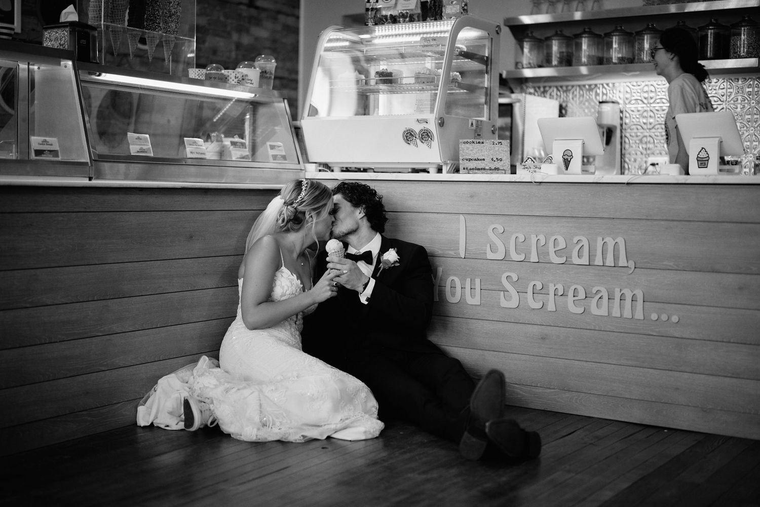 A romantic couple sits on the floor against an ice cream shop counter in black and white photography.