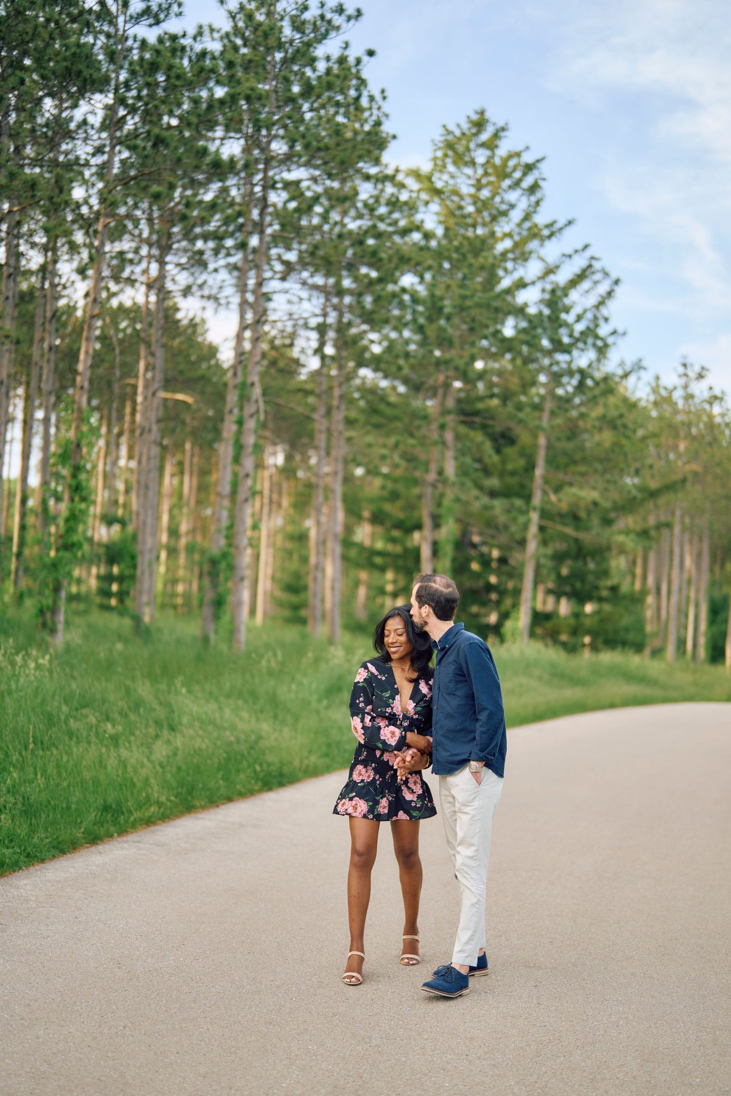 A couple takes a romantic stroll together on a paved path through a scenic pine forest on a sunny day.