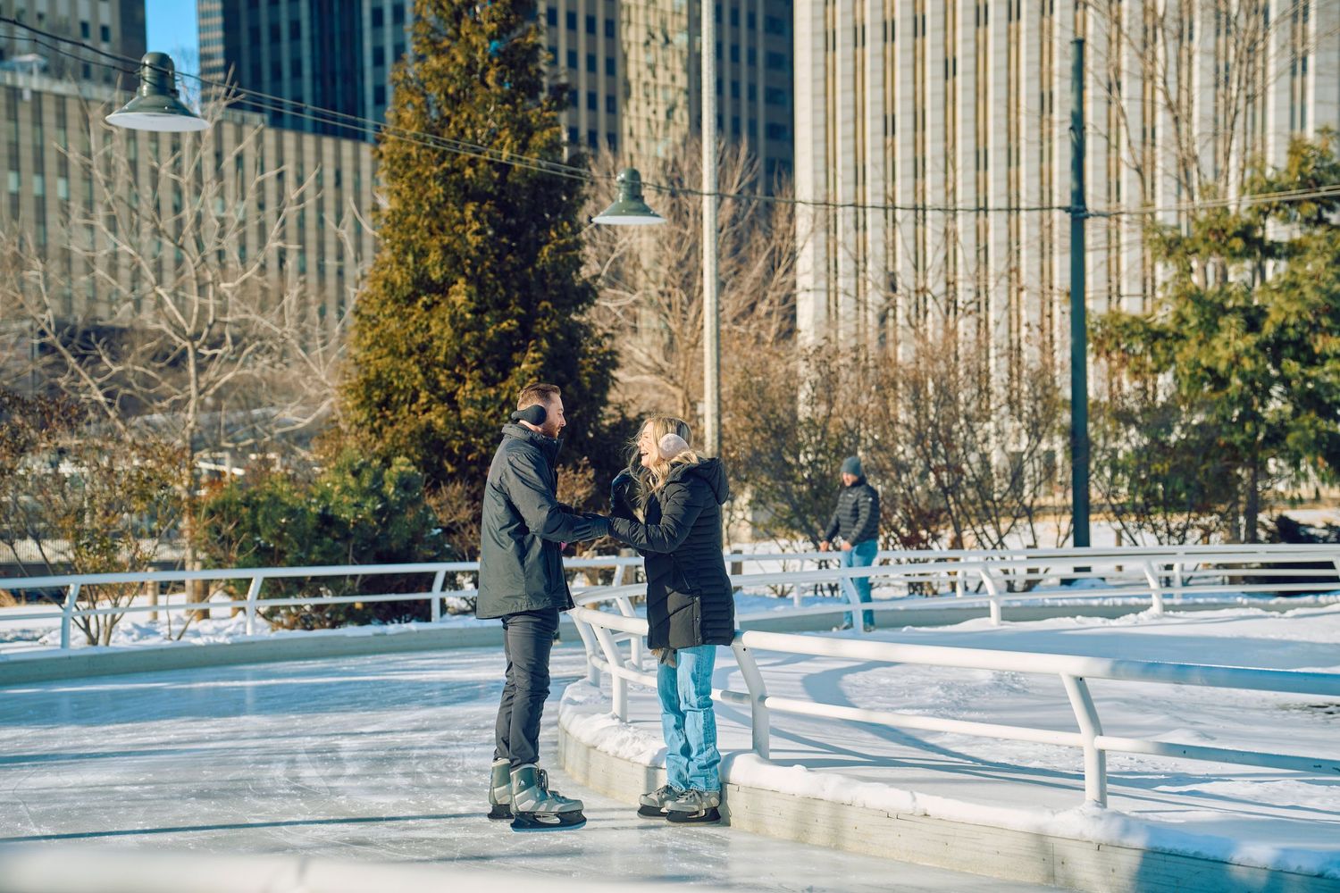 Two people ice skating together at an outdoor rink with city buildings and trees in the background on a winter day.