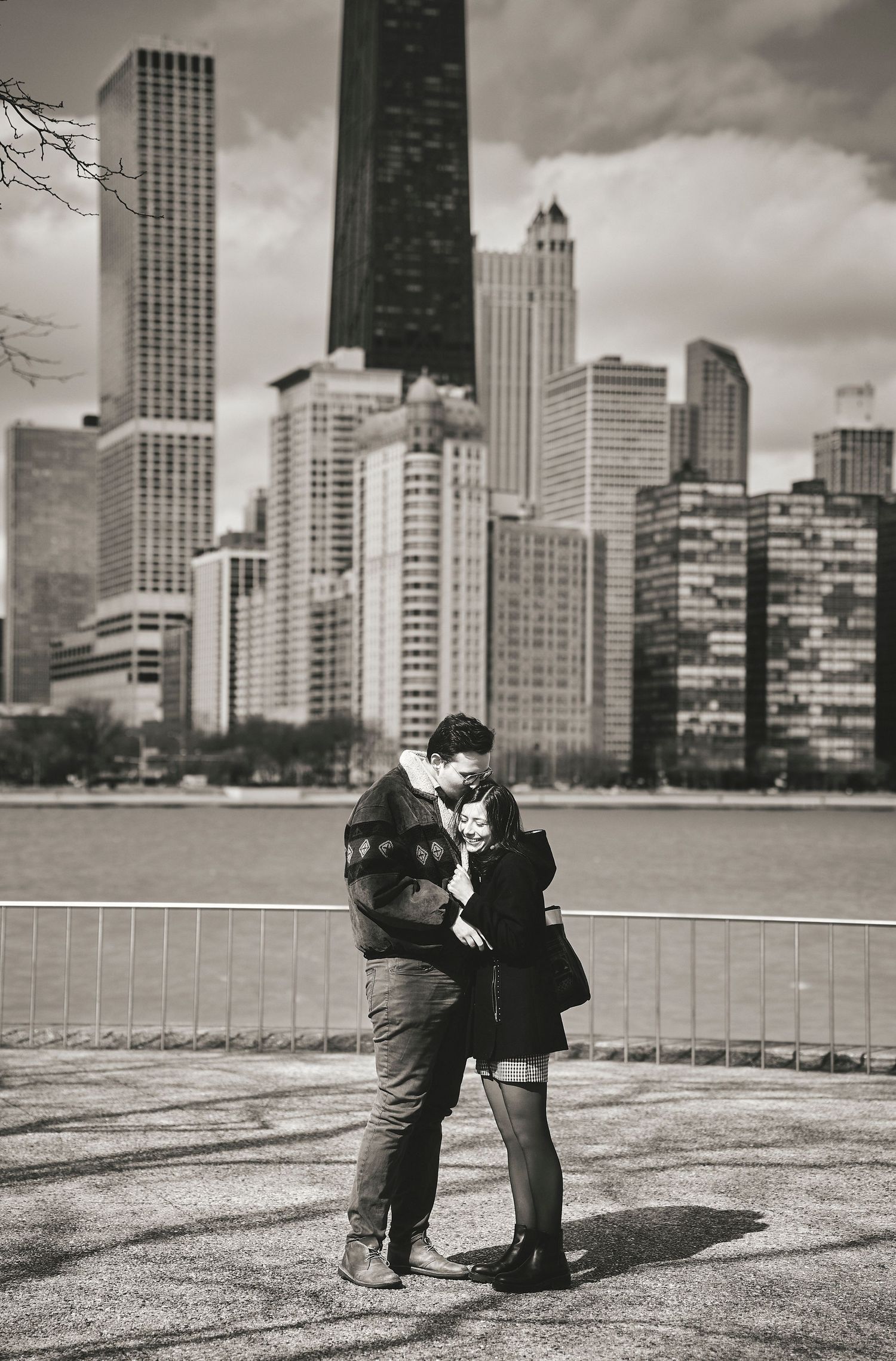 A couple embraces against the dramatic Chicago skyline with skyscrapers and moody clouds in black and white.