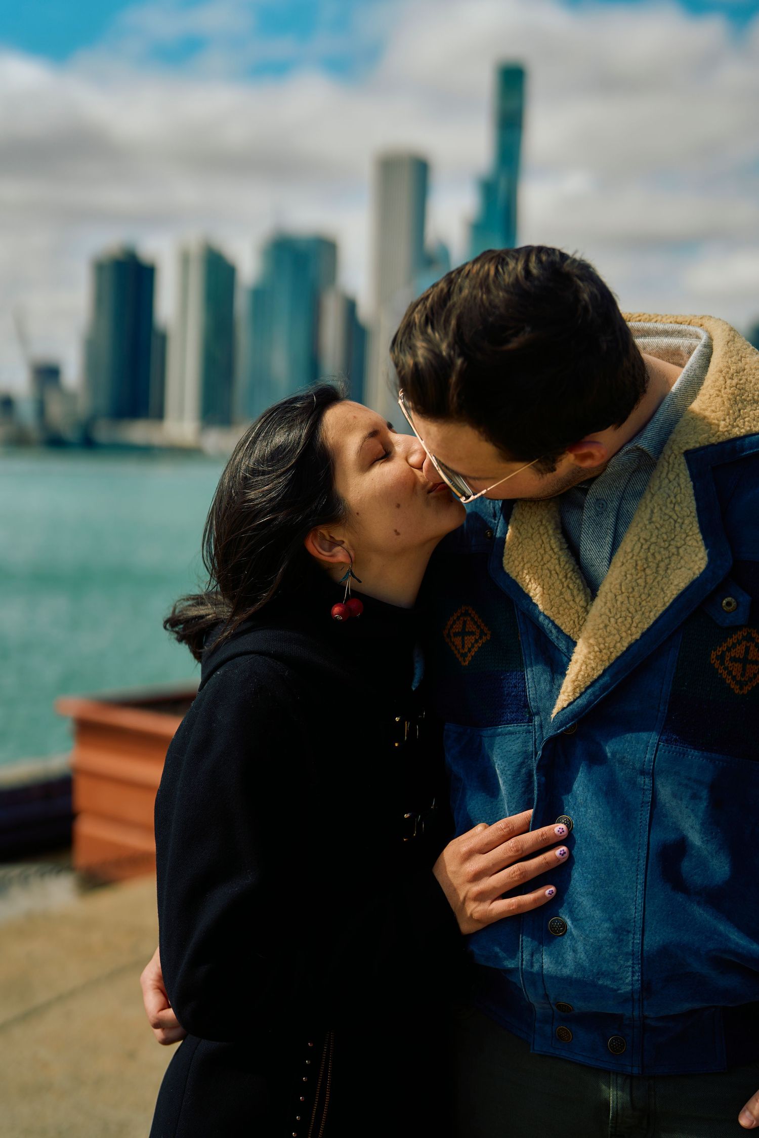 A couple share an intimate kiss along the waterfront with a city skyline of tall buildings in the background.