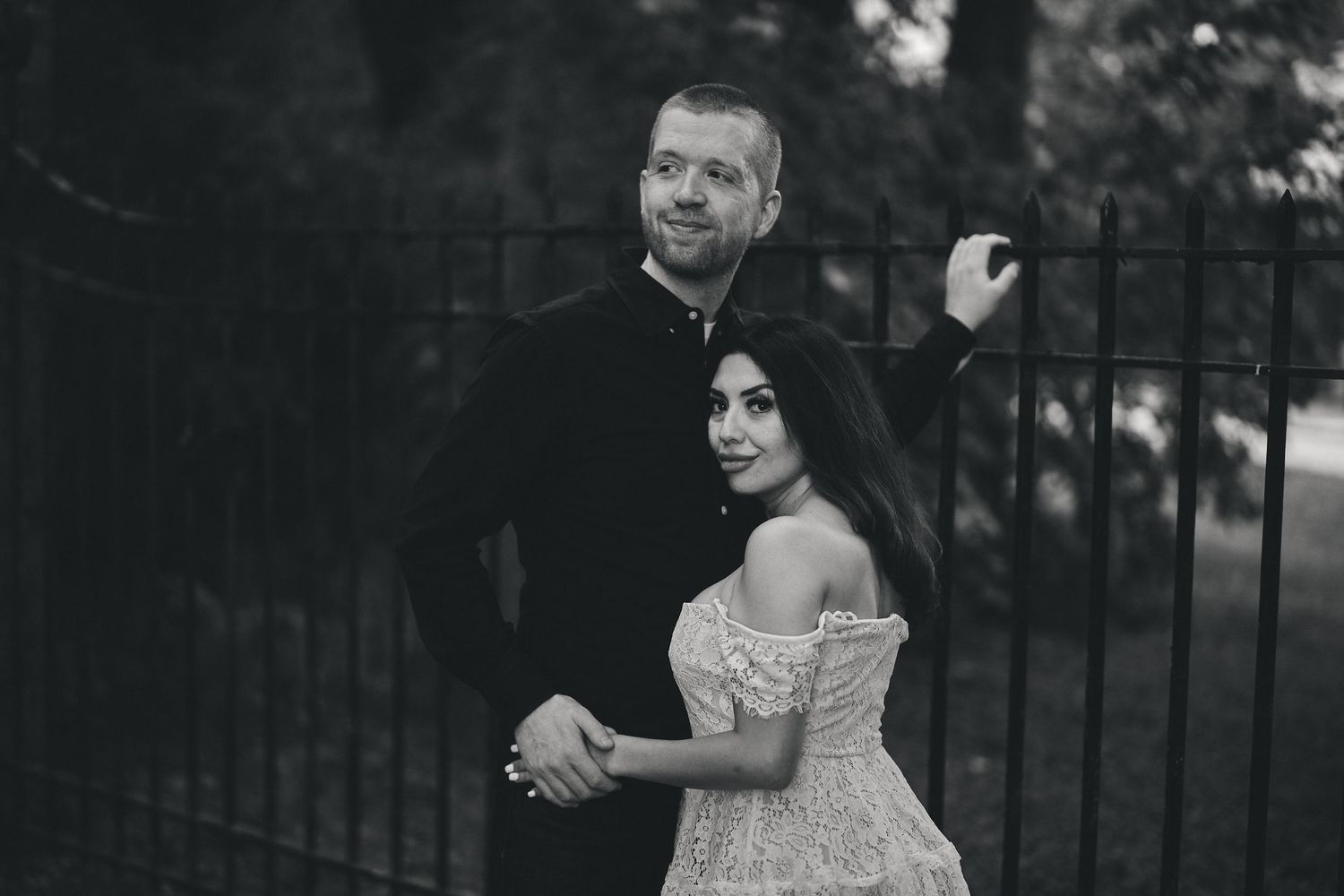Black and white portrait of a couple in elegant wedding attire standing close together by iron gates.