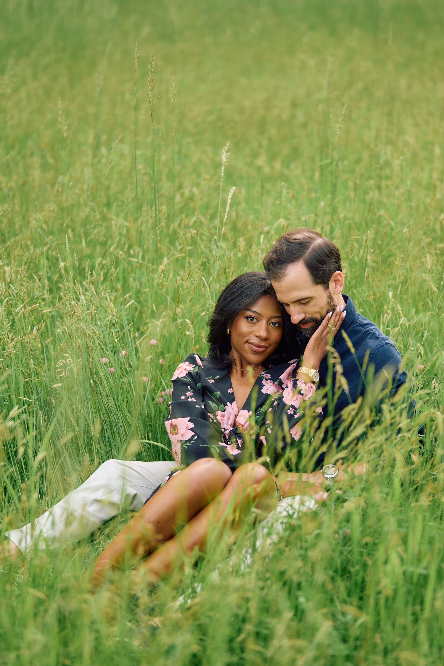 A romantic couple sits close together in a tall grass meadow on a sunny afternoon.