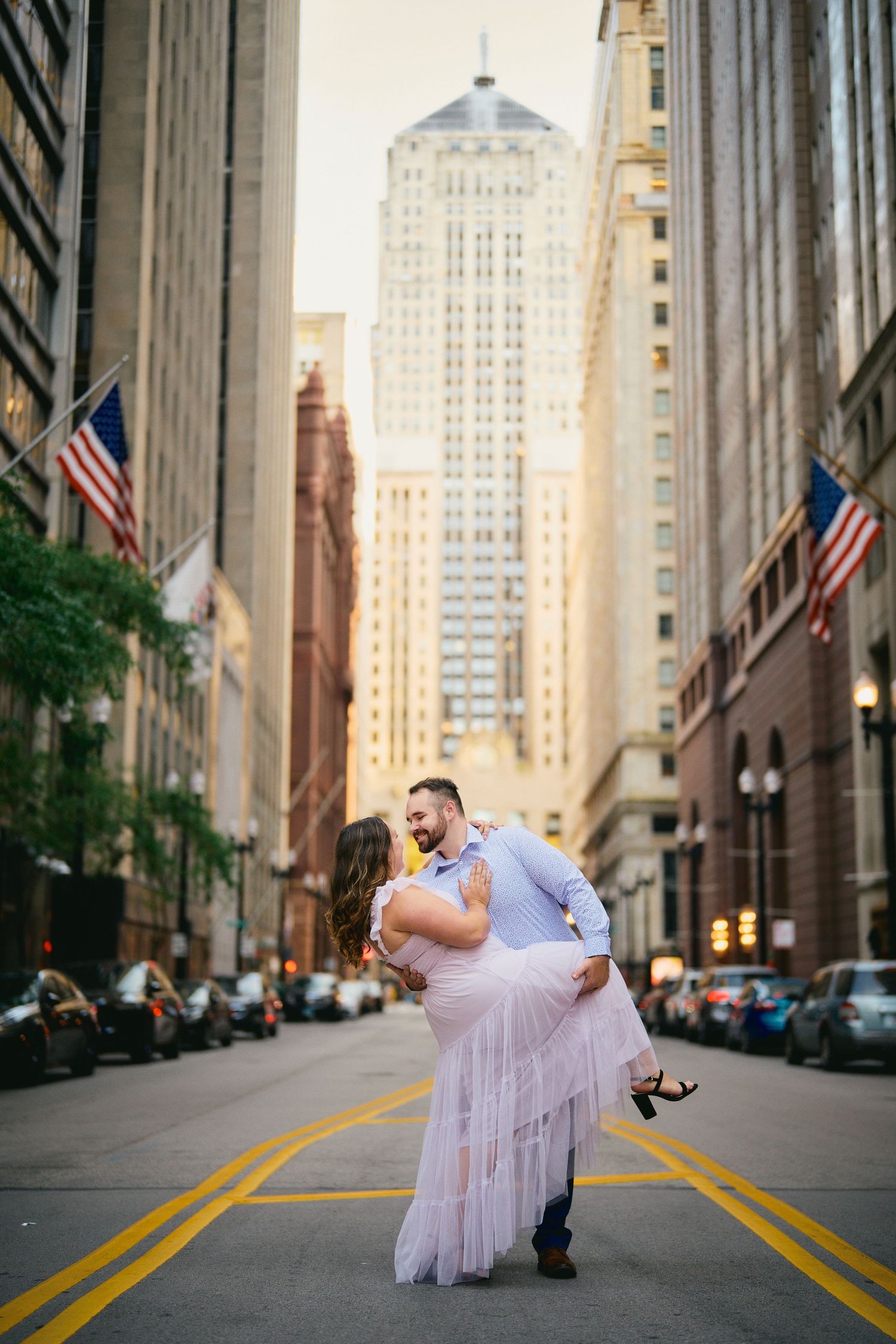 Couple embraces in the middle of a Chicago street with tall buildings and American flags in the background.