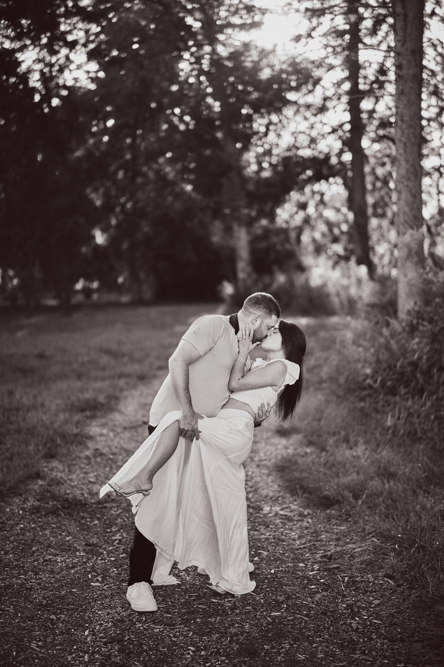 A romantic black and white photograph of a couple embracing and kissing on a tree-lined path.
