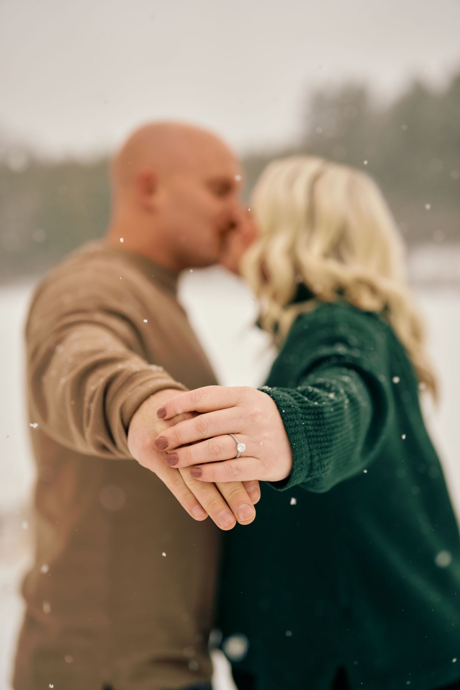 A couple sharing a romantic kiss in the snow while showing off an engagement ring.
