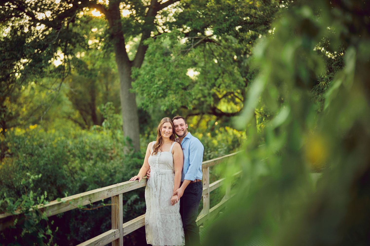 Couple stands together on wooden bridge surrounded by lush green foliage in romantic outdoor setting.