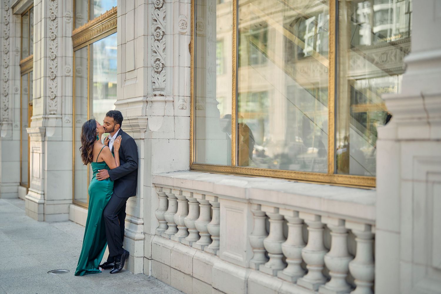 A couple in formal attire embraces against an ornate stone building facade with tall windows and decorative balustrades.