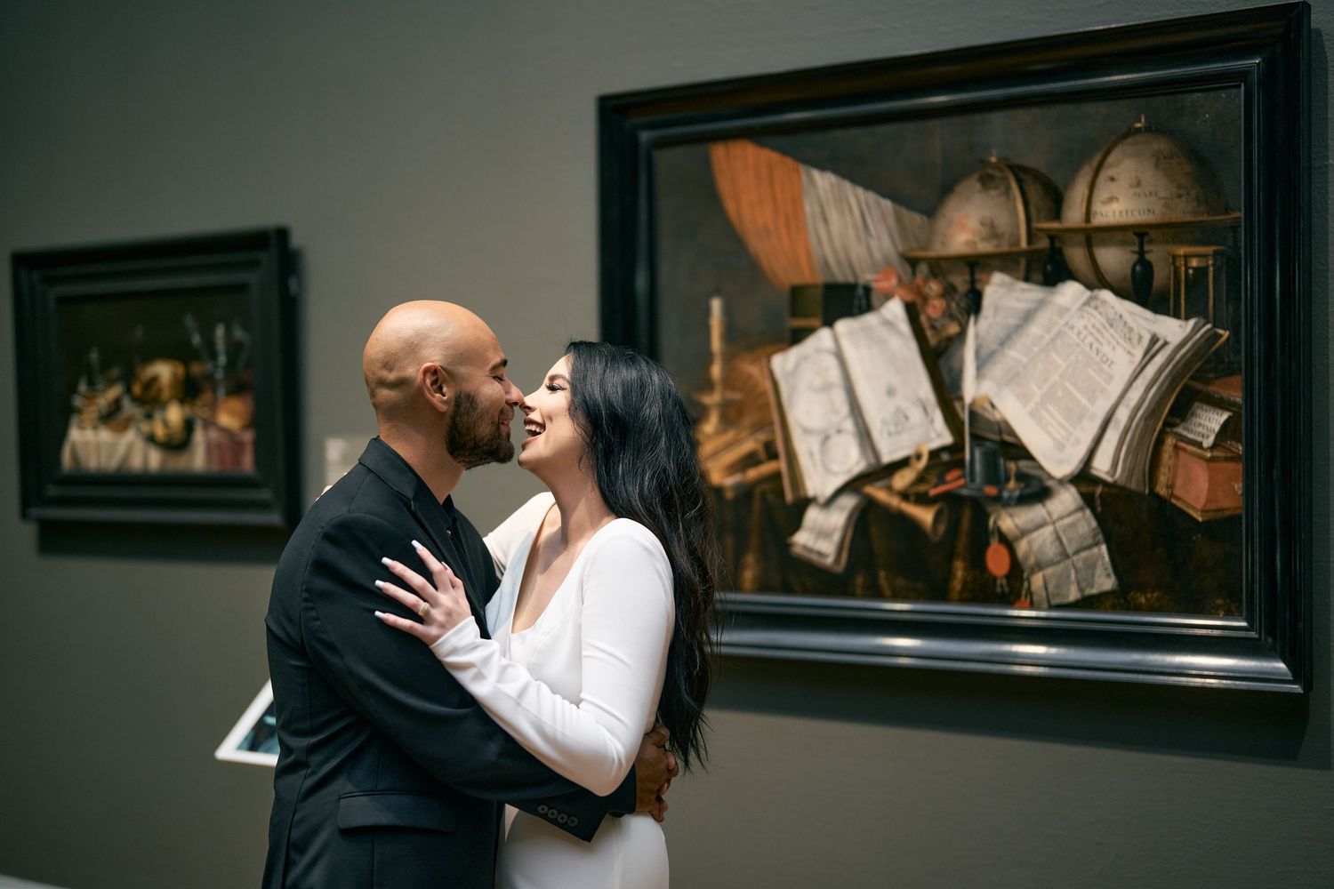 A couple shares a tender moment while embracing in front of classical artwork displayed in a museum gallery.