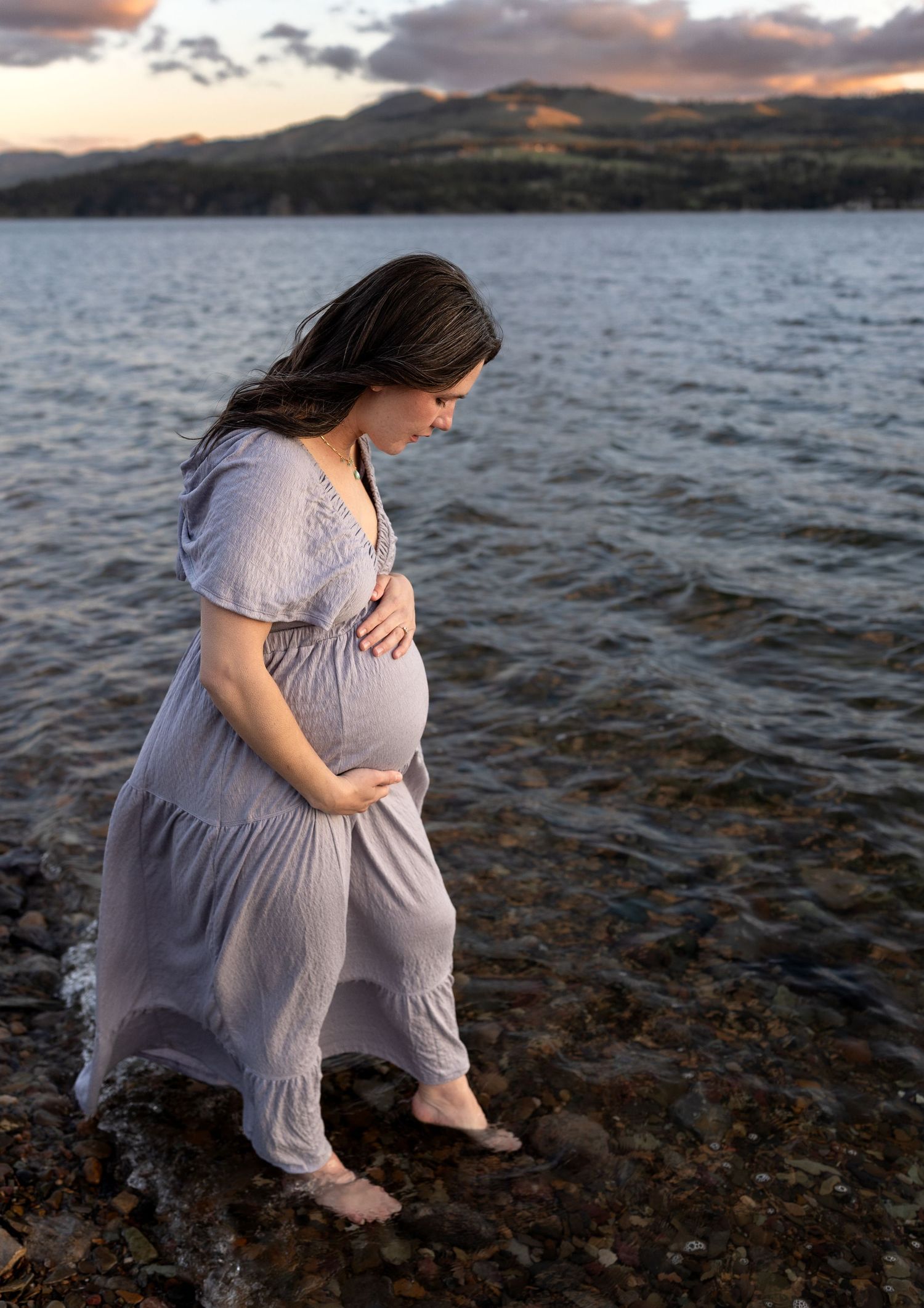 A pregnant woman stands ankle deep at the shore of a lake as she caresses her belly