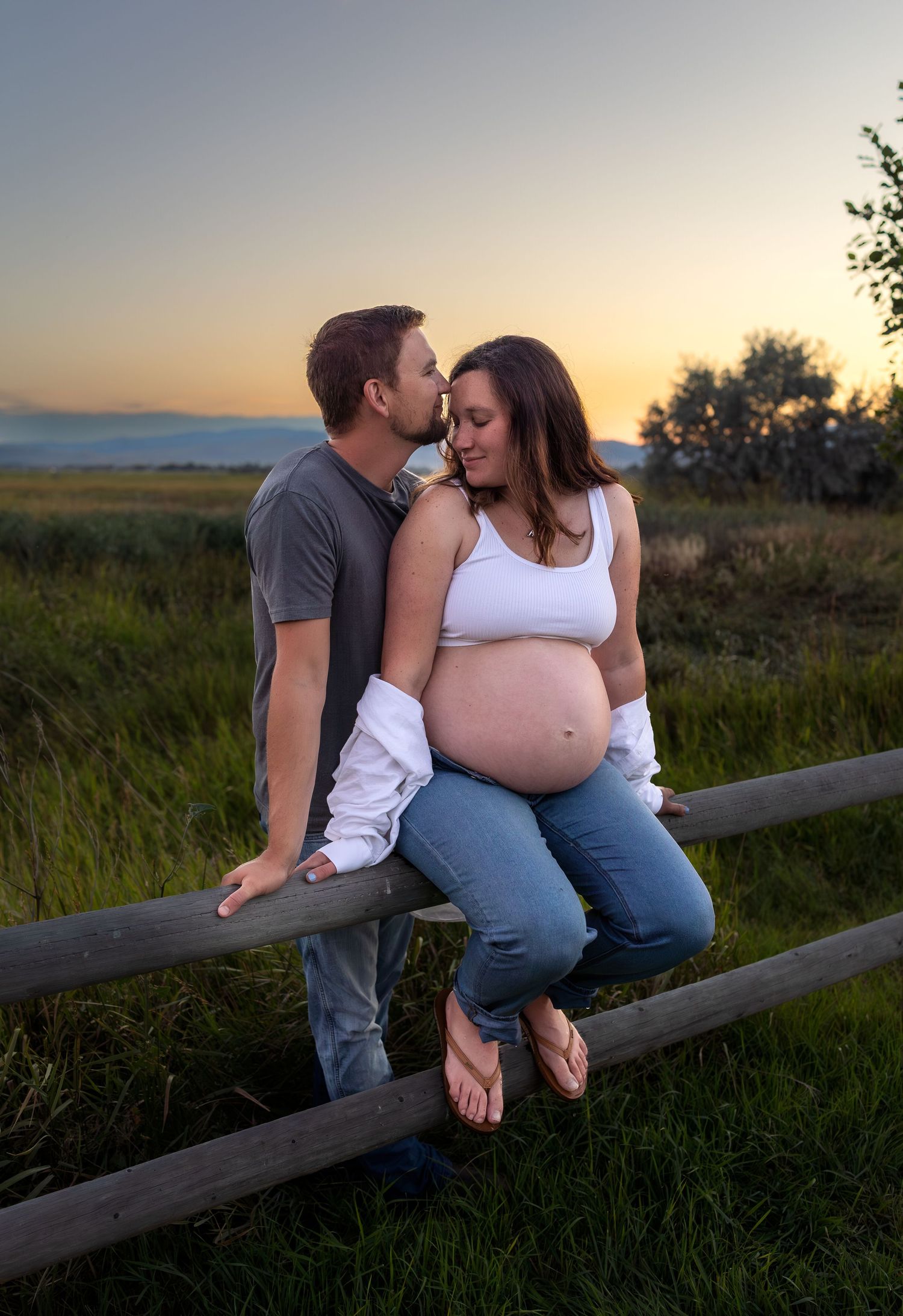 A pregnant woman sits on a wood fence while her husband stands behind her and kisses her forehead