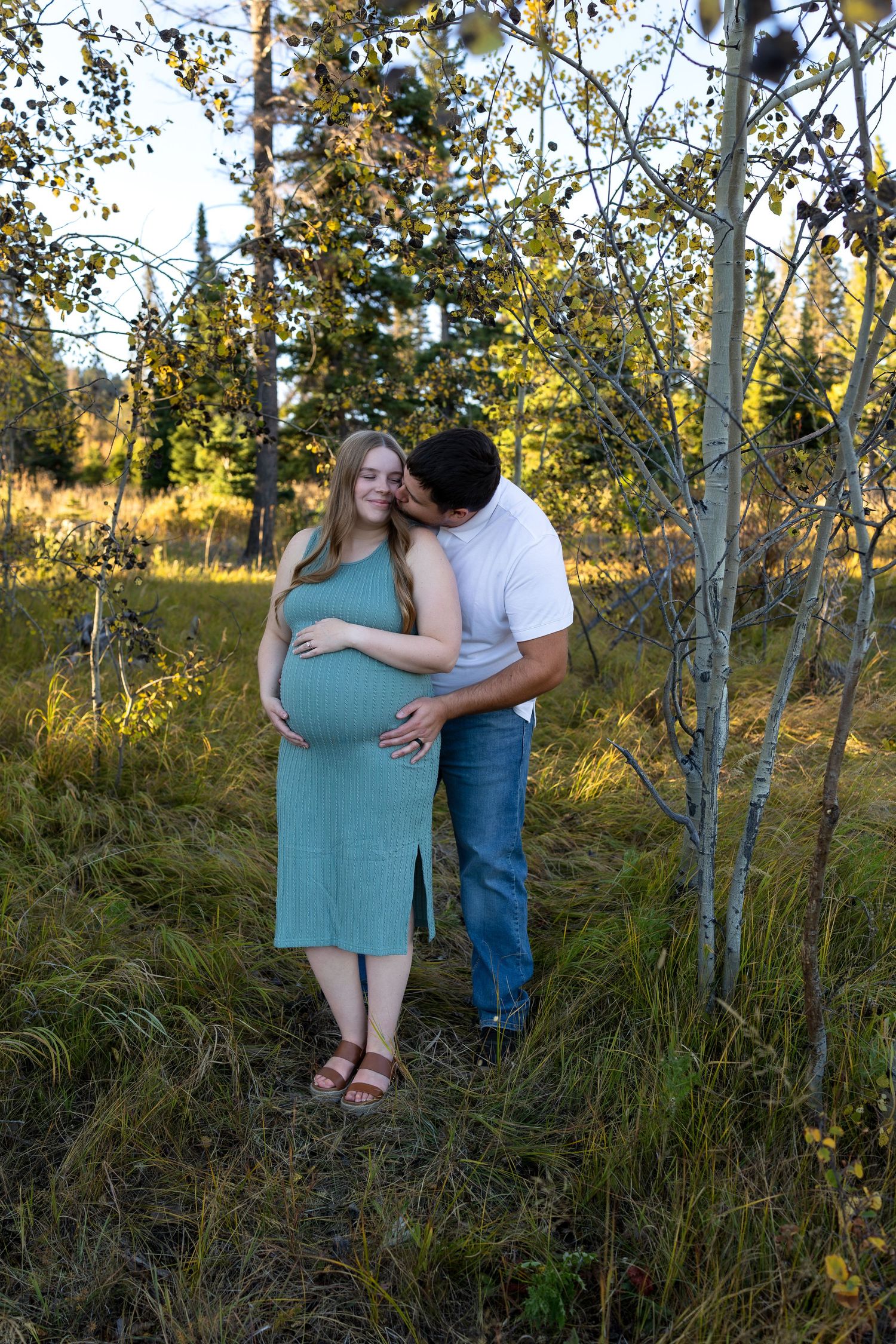 A pregnant woman stands in front of her husband as he leans around to kiss her cheek