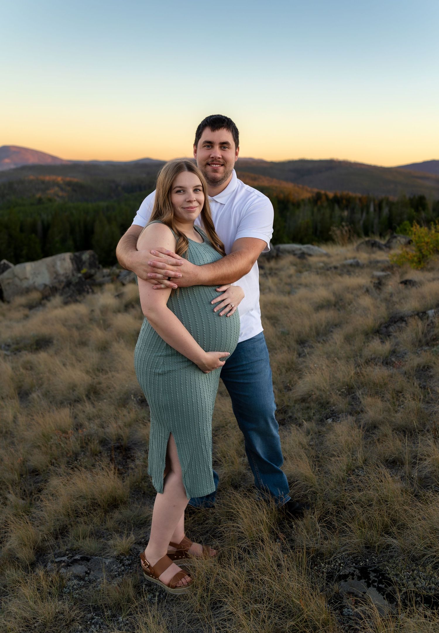A pregnant woman smiles at the camera while her husband stands behind her and embraces her