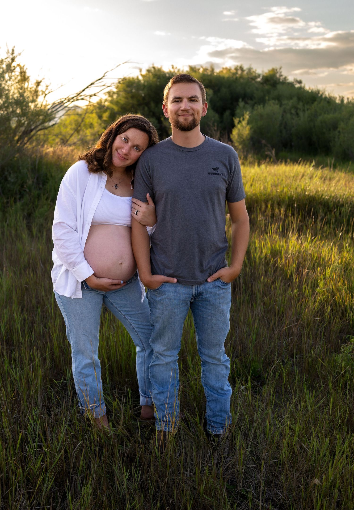 A pregnant woman leans her head on her husbands shoulder in a green field