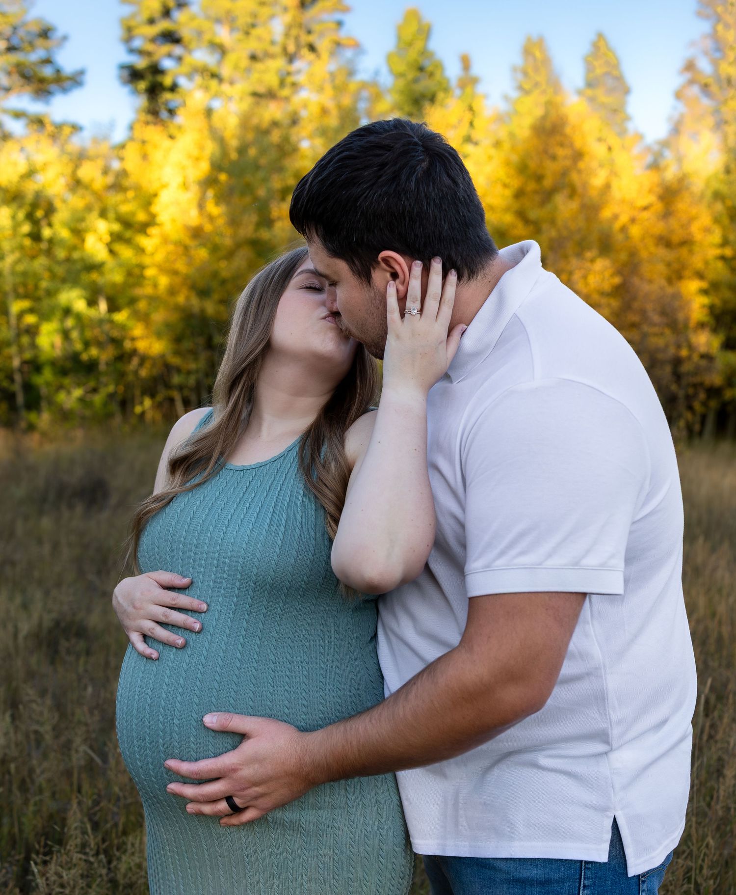 A pregnant woman and her husband share a kiss in a field with autumn trees behind them