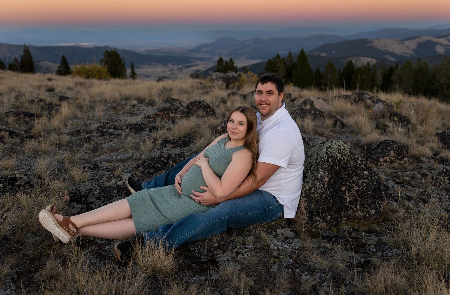 A man and pregnant woman sit on top of a mountain at sunset