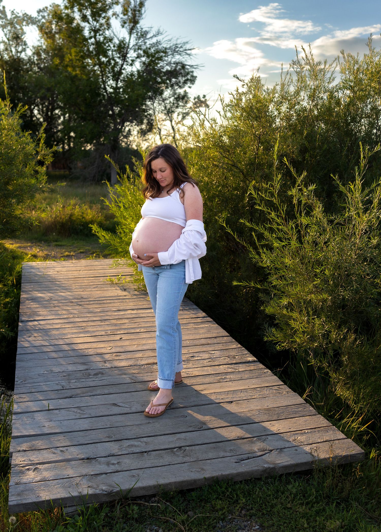 A pregnant woman stands on a wood bridge with trees behind her