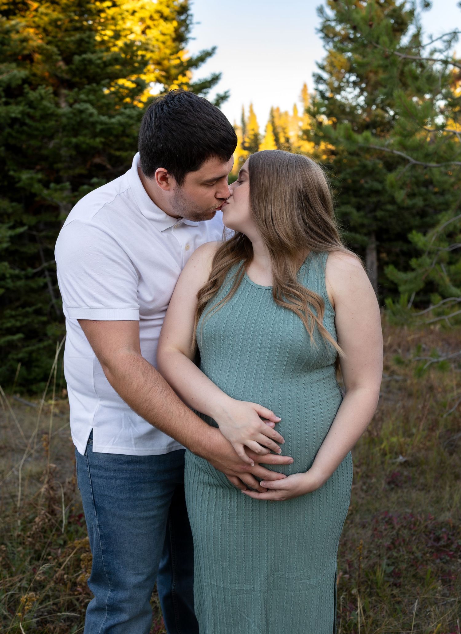 A pregnant woman stands in front of her husband as he leans around her for a kiss