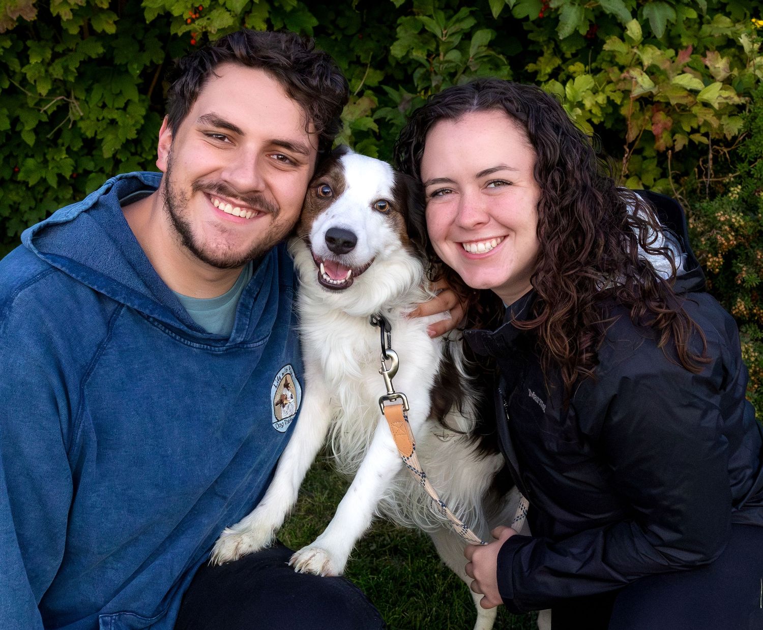 A man and woman pose with their dog between them in front of a green hedge