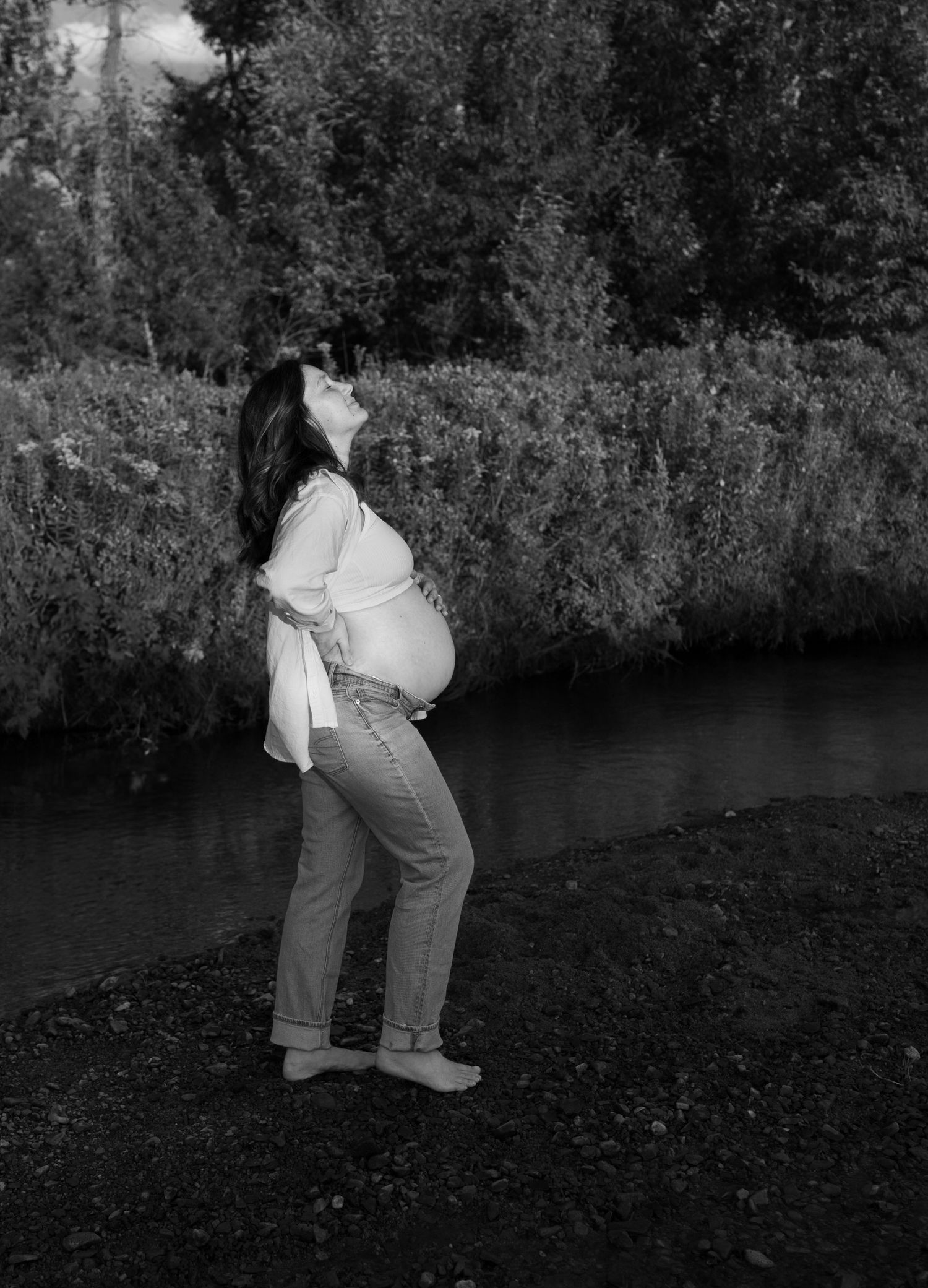A black and white image of a pregnant woman at the bank of a river, she leans her face up to the sun with her eyes closed