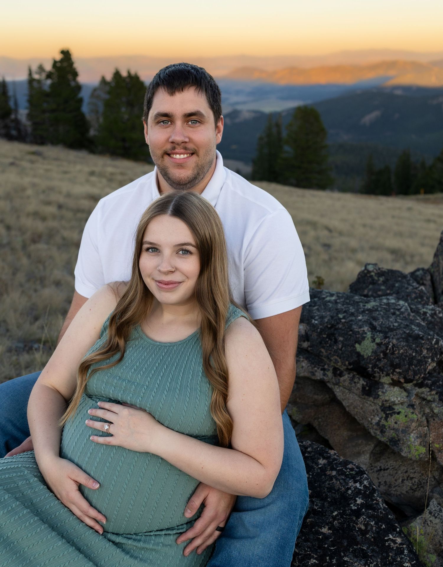 A pregnant woman sits between her husbands legs as she caresses her belly, a mountain view behind them