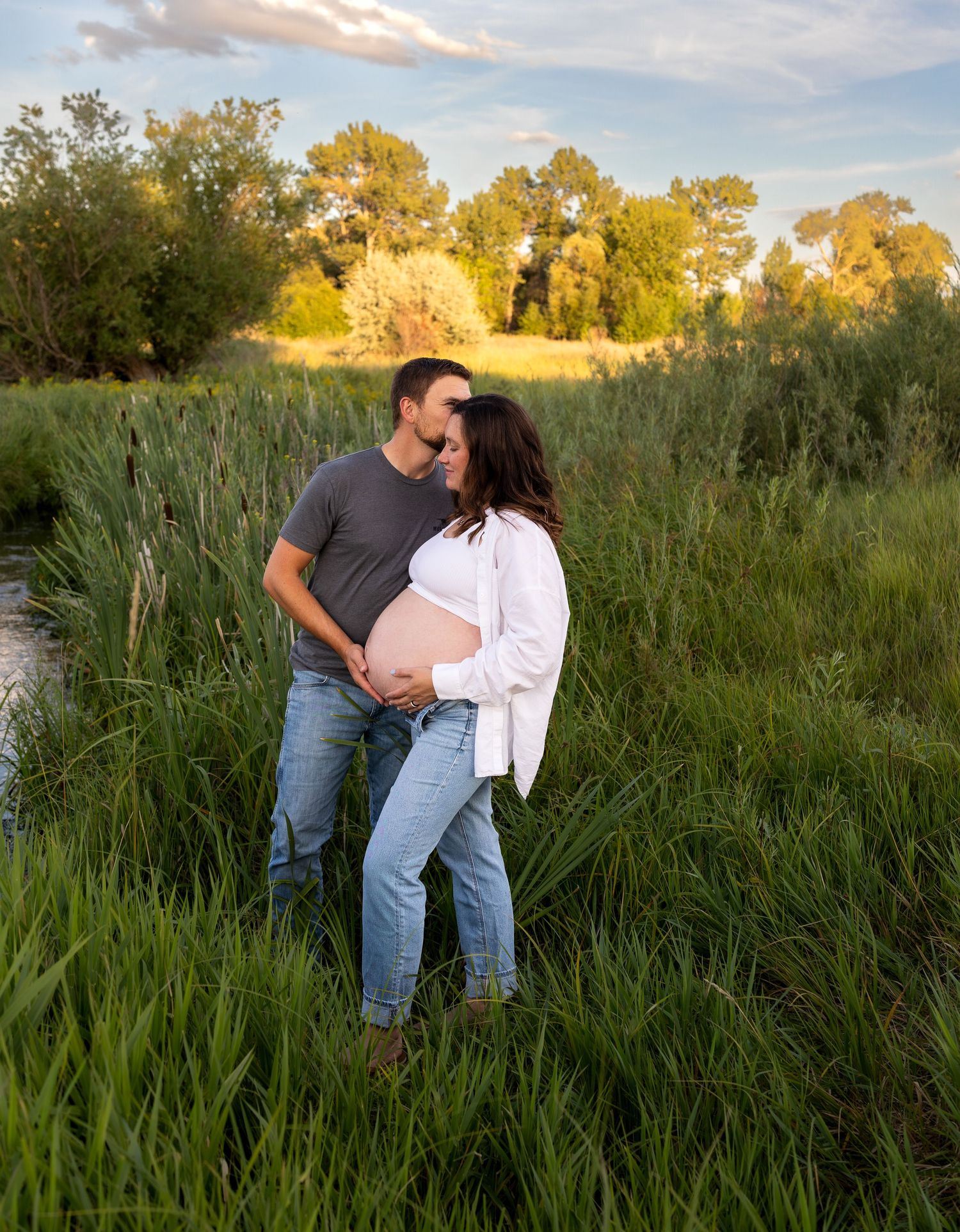 A man kisses his wife's head as he caresses her pregnant belly in a green field