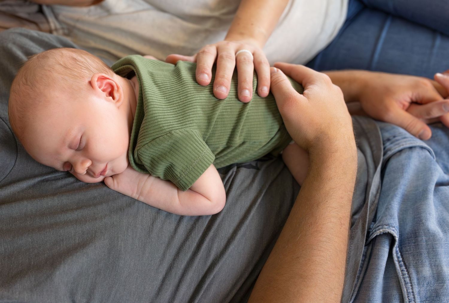 A close up image of a newborn baby boy sleeping on his Dad's chest, with his Mom's hand resting on his back.