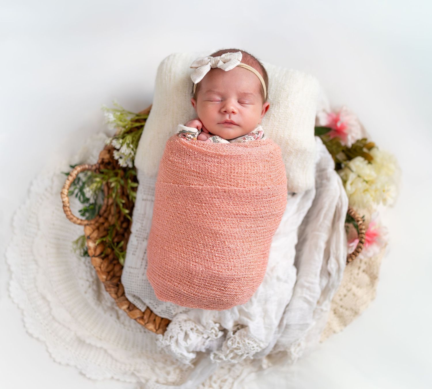 A posed image of a newborn girl in a pink wrap with a bow on her head.