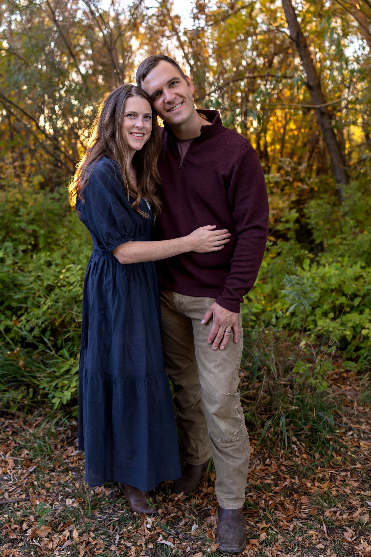 a husband and wife embrace in front of trees with autumn colors at sunset
