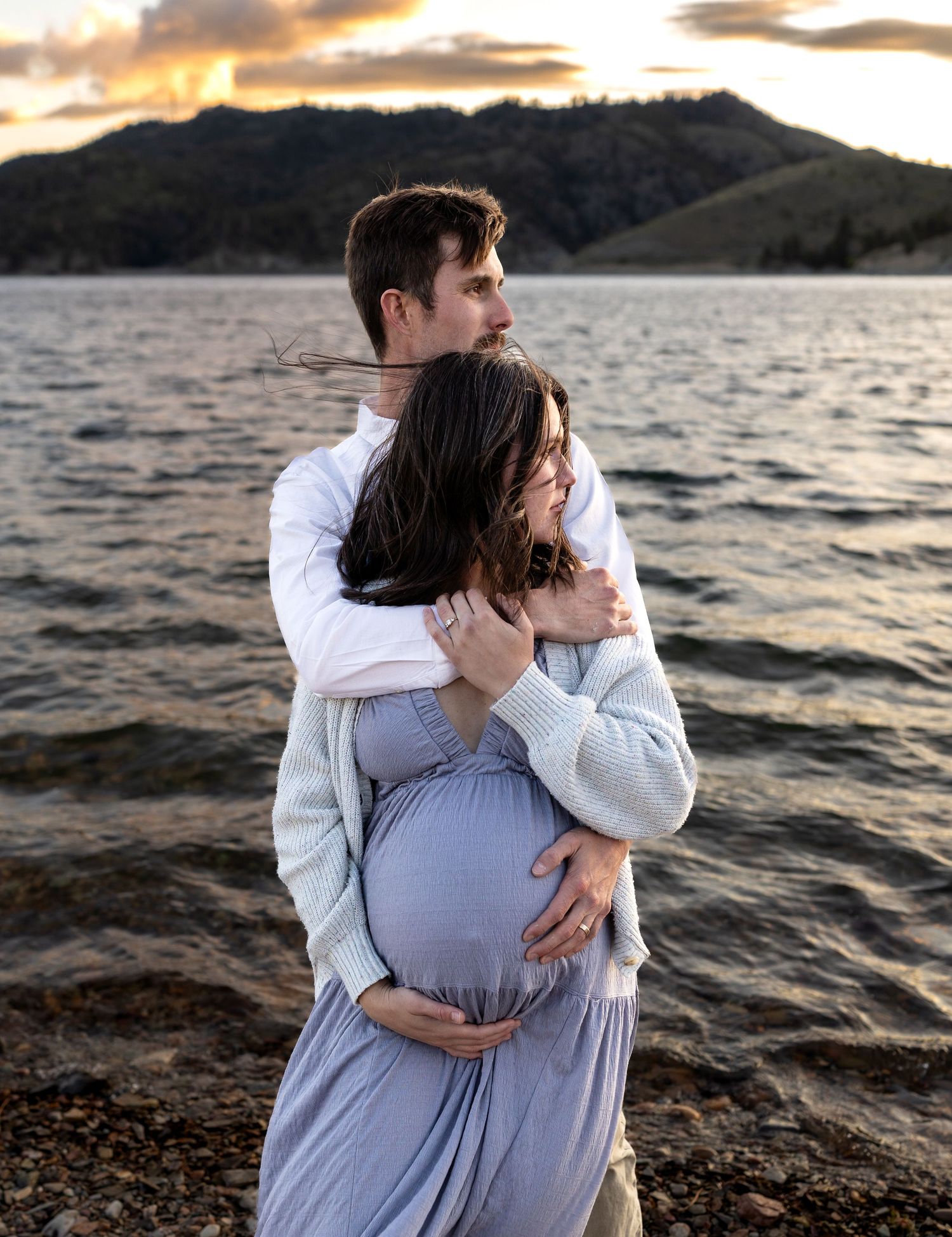 A pregnant woman and her husband pose in front of a lake at sunset