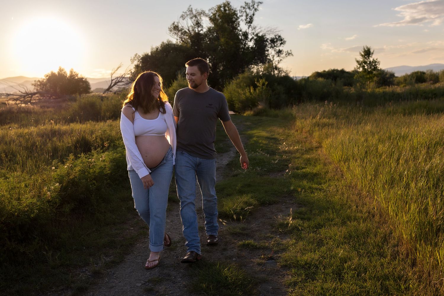 A pregnant woman and her husband walk and laugh down a country lane surrounded by greenery
