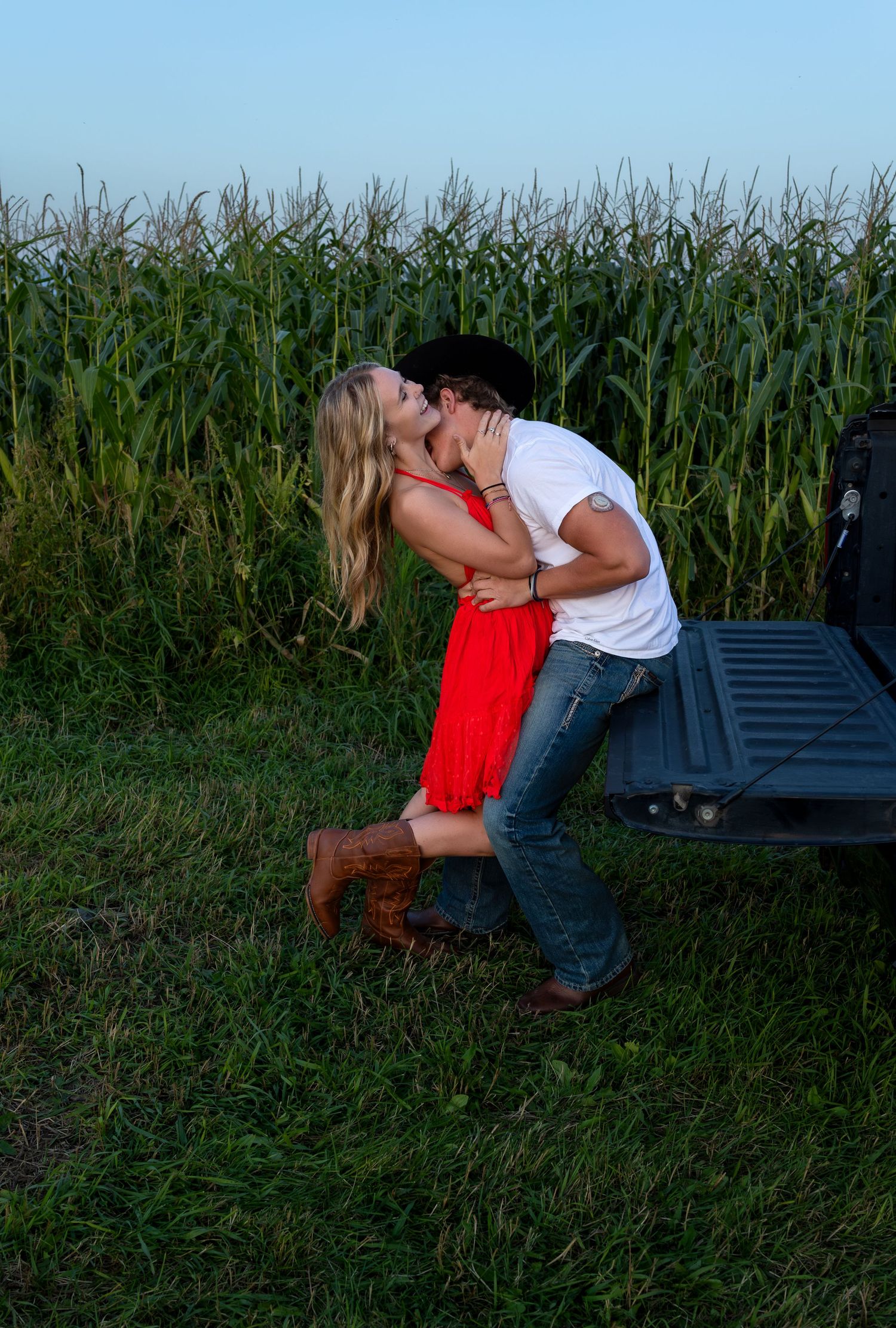 A young man sits on the tailgate of a truck as his holds his girlfriend close and kisses her neck