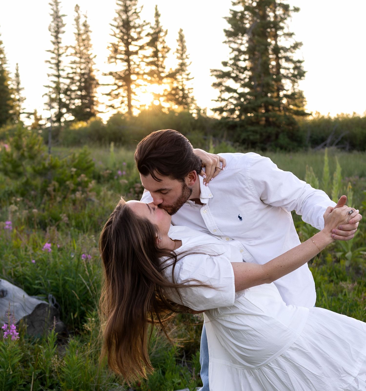 A man dips his fiancé in front of grassy flower filled meadow