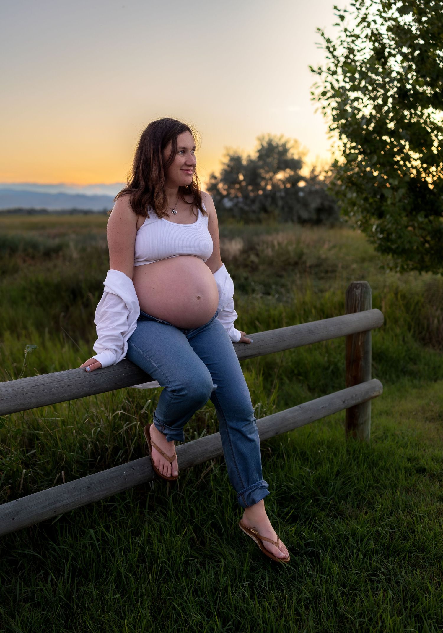 A pregnant woman sits on top of a wood fence in a field at sunset