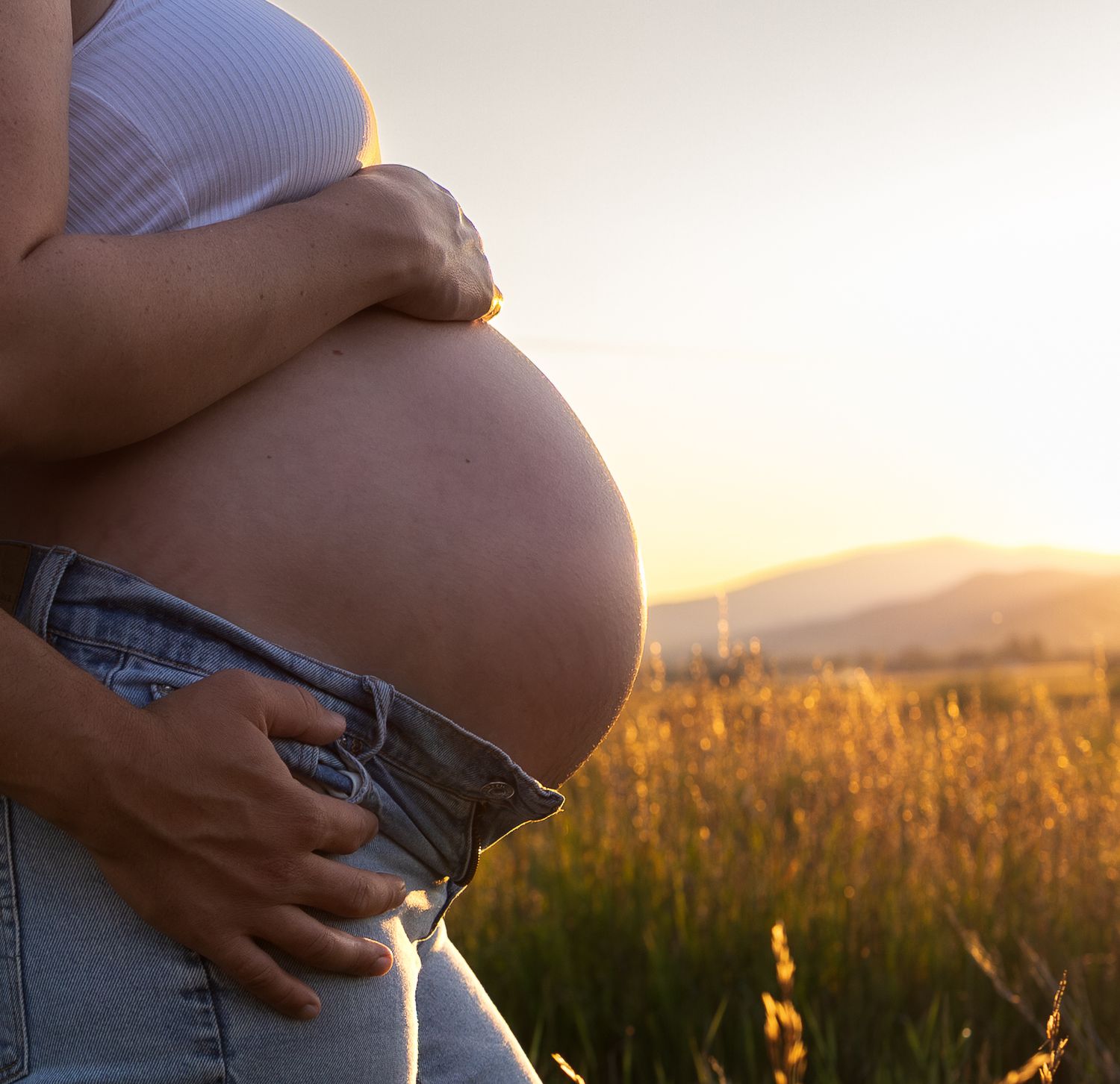 An up close image of a pregnant belly, husbands hand on her hip, sun shining on the field behind them