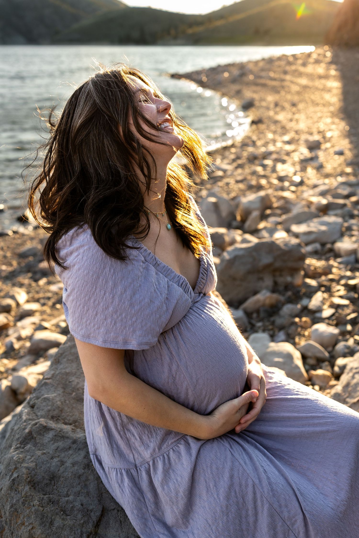 A pregnant woman laughs joyfully in front of a lake while holding her belly