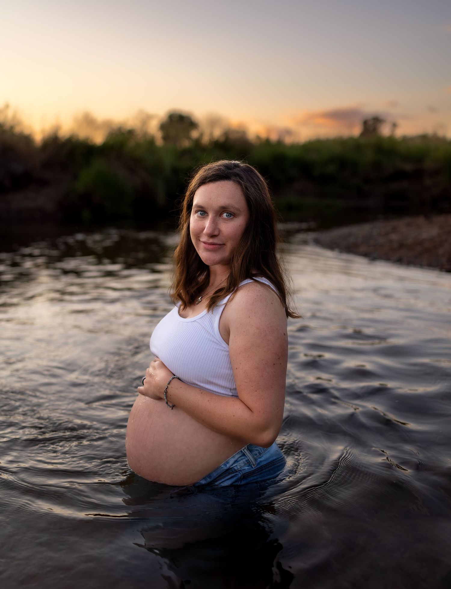 A pregnant woman poses half submerged in a river at sunset, with her belly peeking out