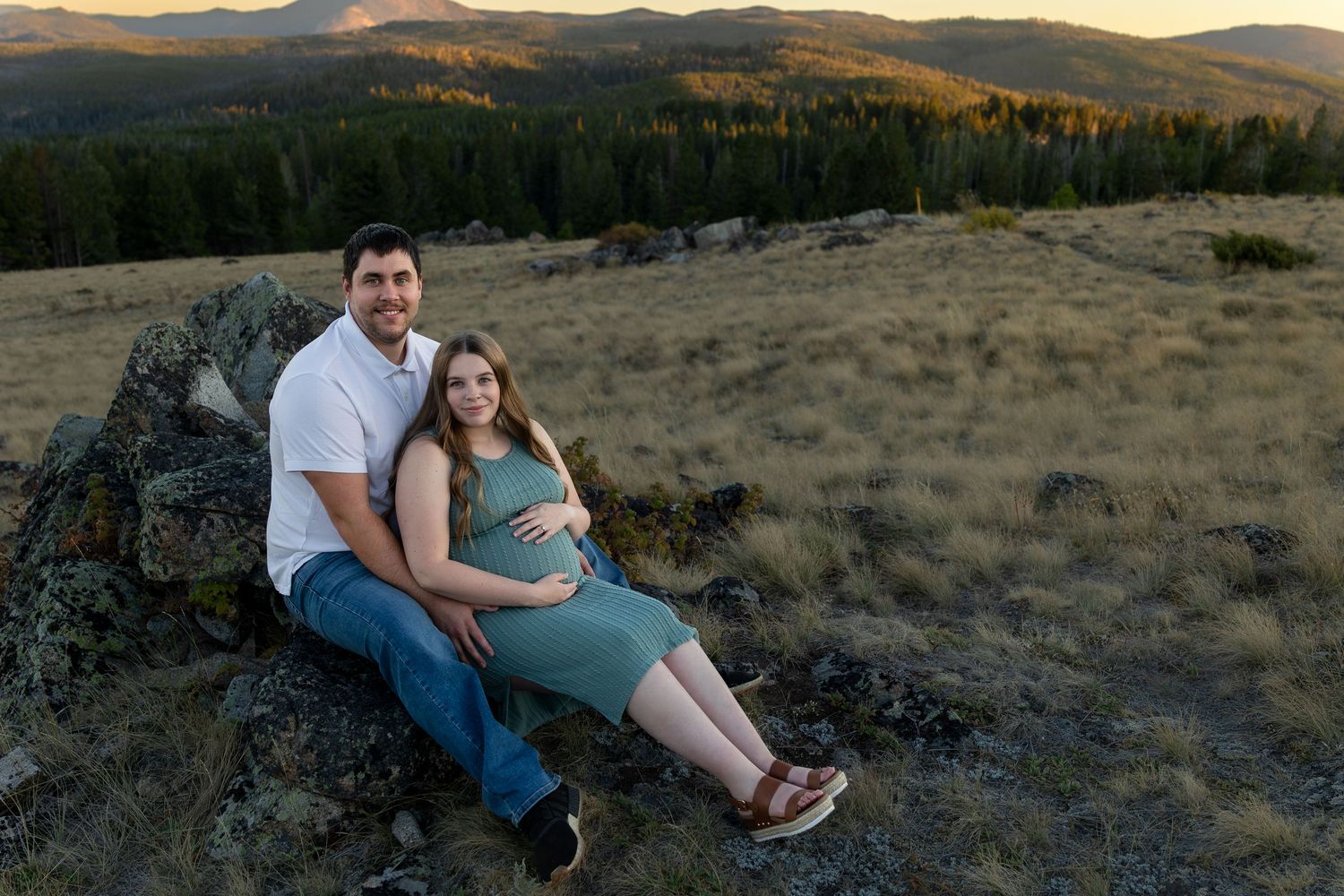 A pregnant woman and her husband sit and lean against a big boulder on top of a mountain