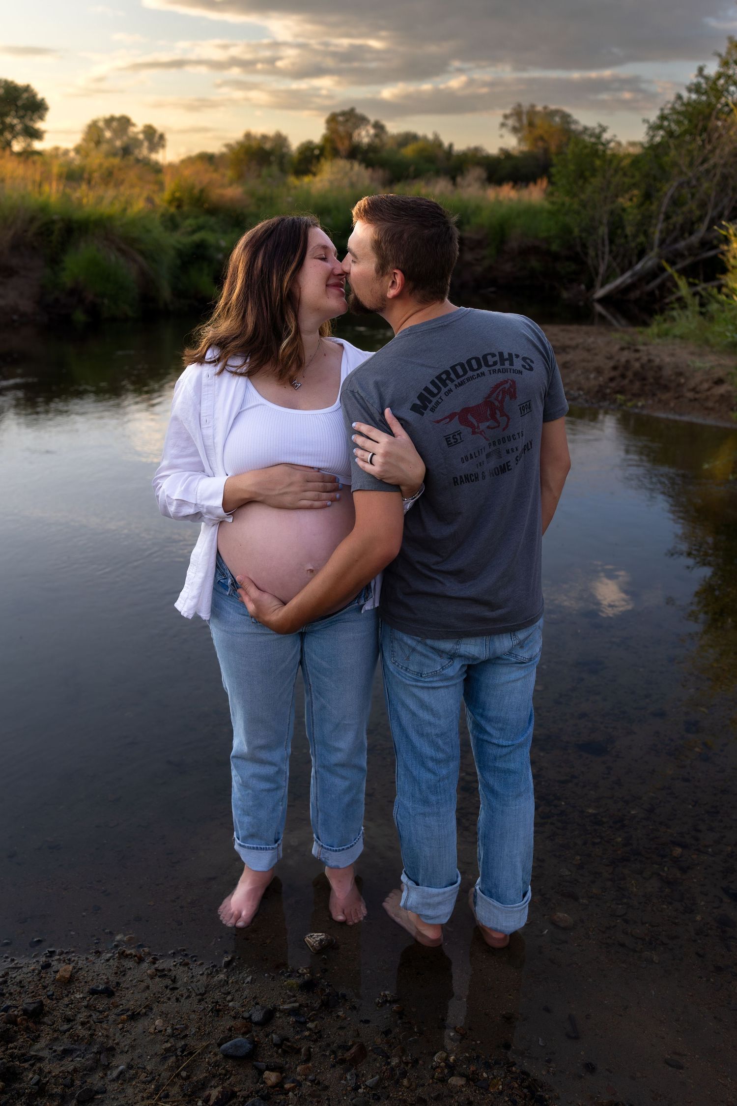 A pregnant woman and her husband share a kiss standing in a shallow creek