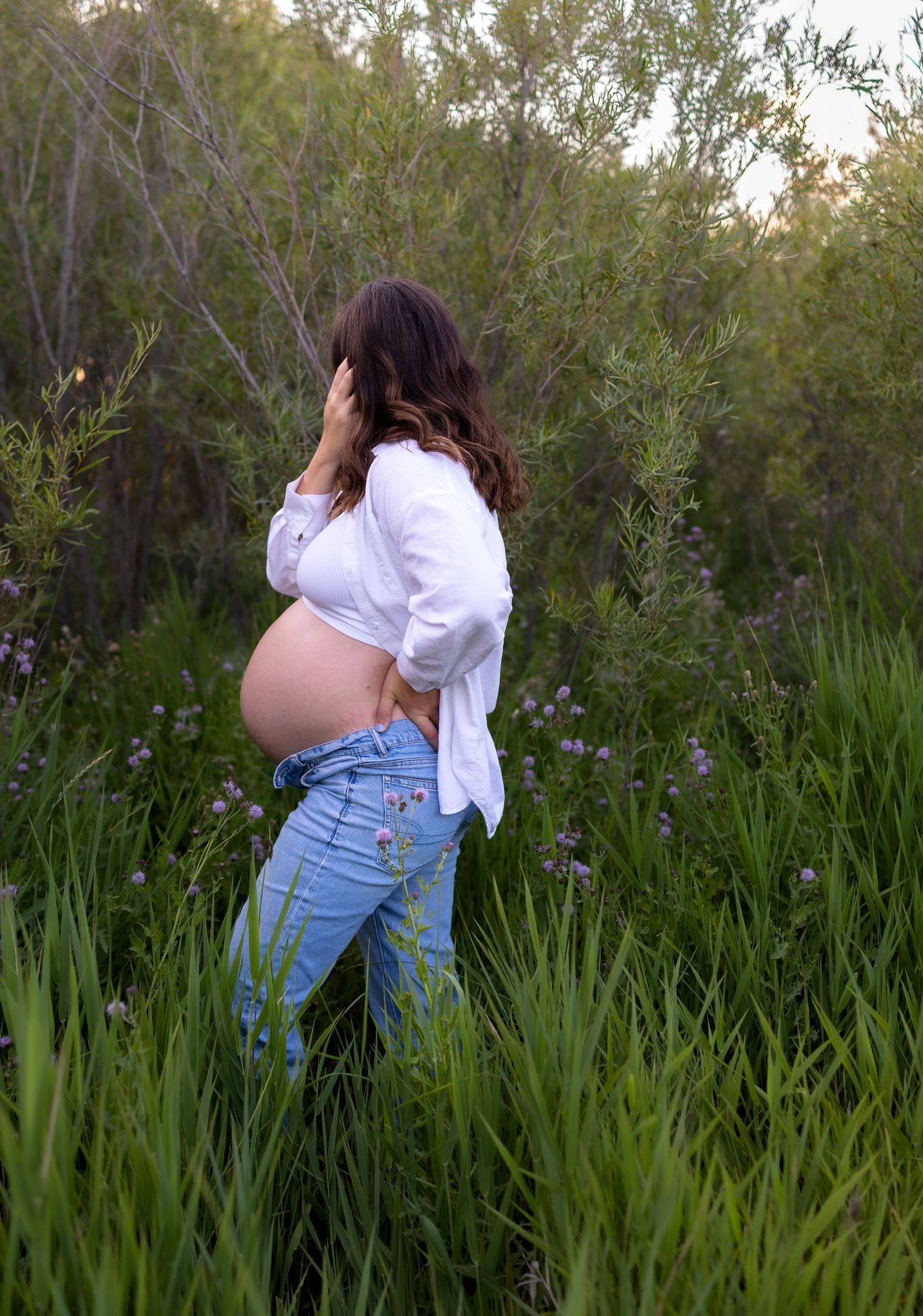 A pregnant woman shows off her belly as she looks away from the camera, she stands in long green grass and trees