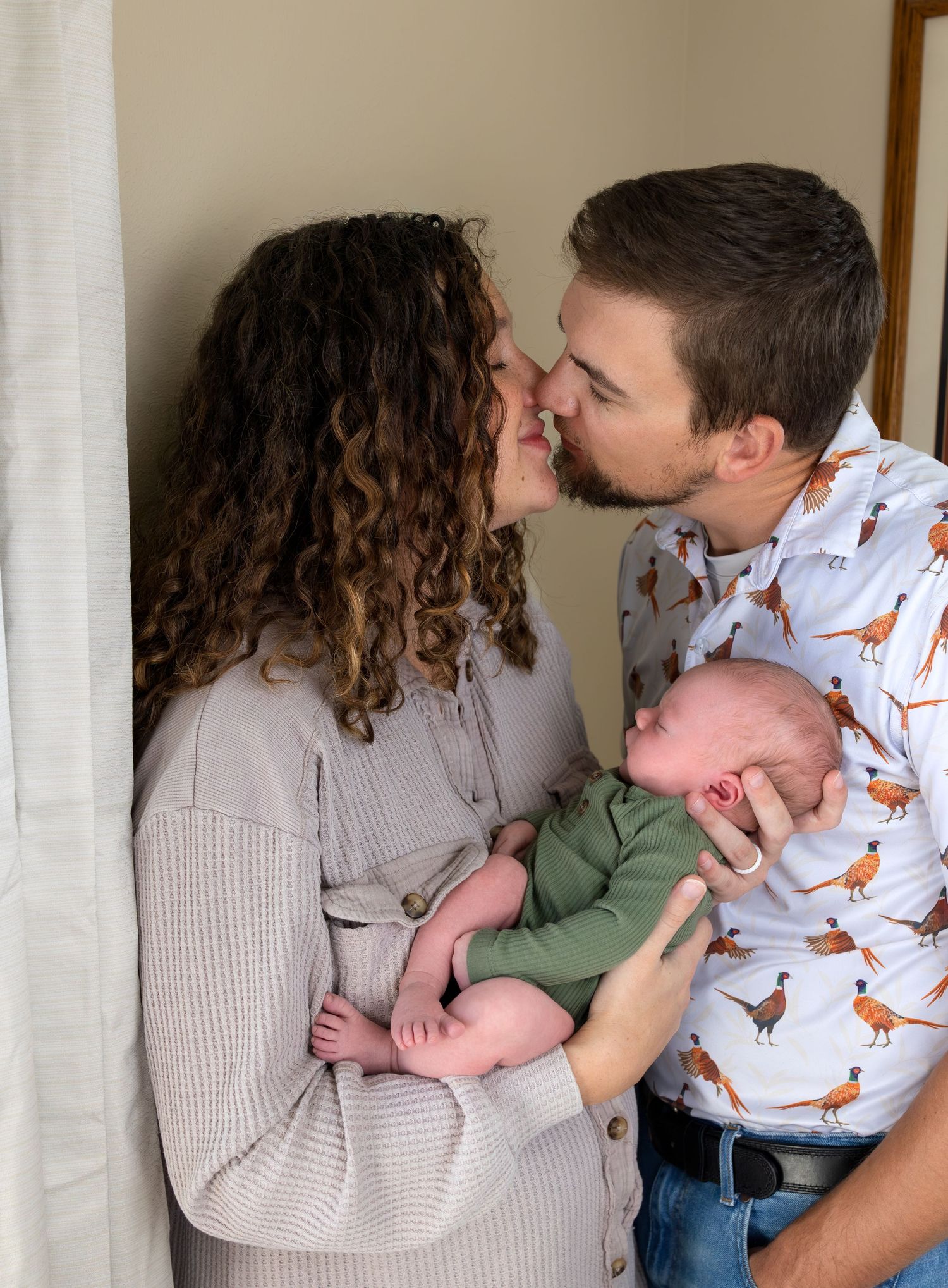 A Mom and Dad share a kiss in their newborn son's nursery, with the newborn baby held in his Mother's arms.