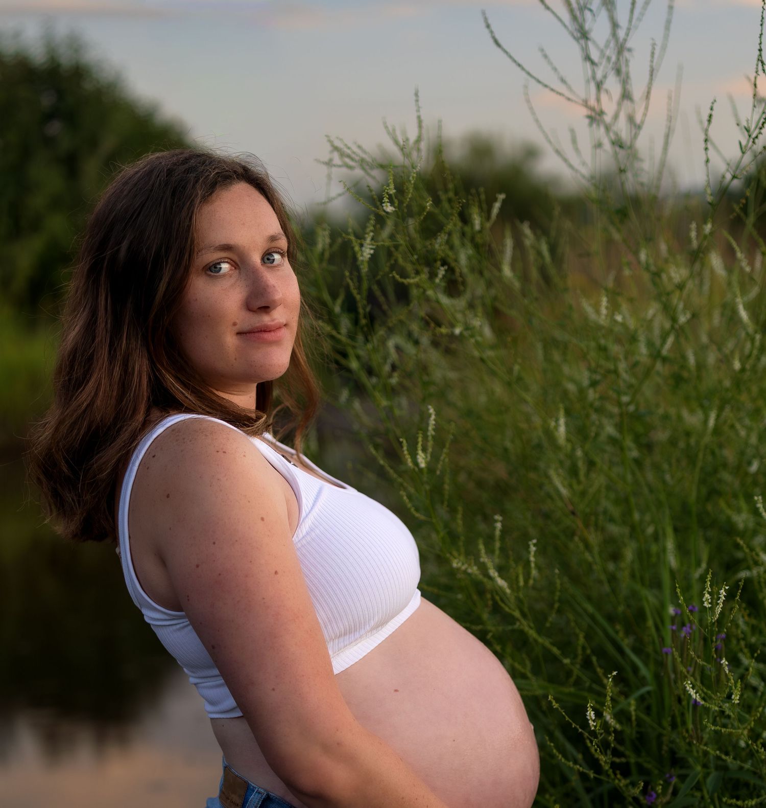 A pregnant woman poses in front of a green bush and river