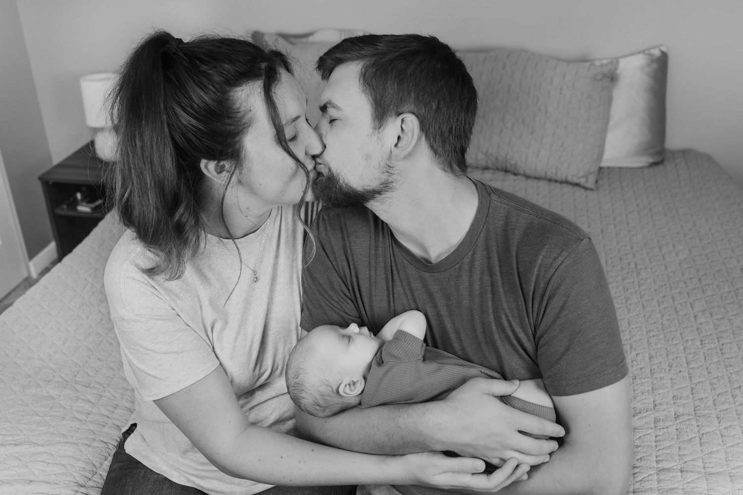 A mom and dad share a kiss on their bed, with their newborn baby boy in their arms.