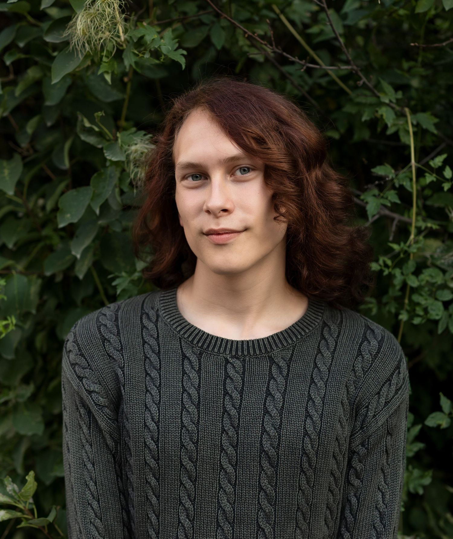 A young man poses in front of a green hedge