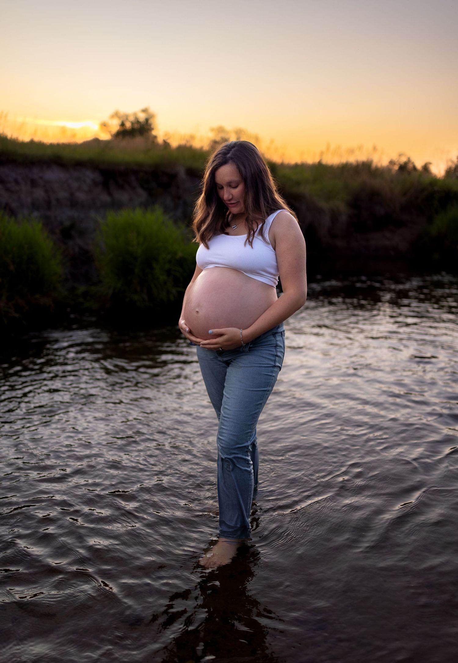A pregnant woman stands in a river at sunset as she caresses her belly